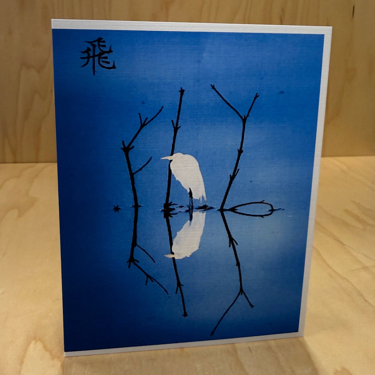 A square art print or card with a blue background, featuring a white bird silhouette perched on dark branch lines and a black Japanese character in the upper left, displayed on a light wood surface.