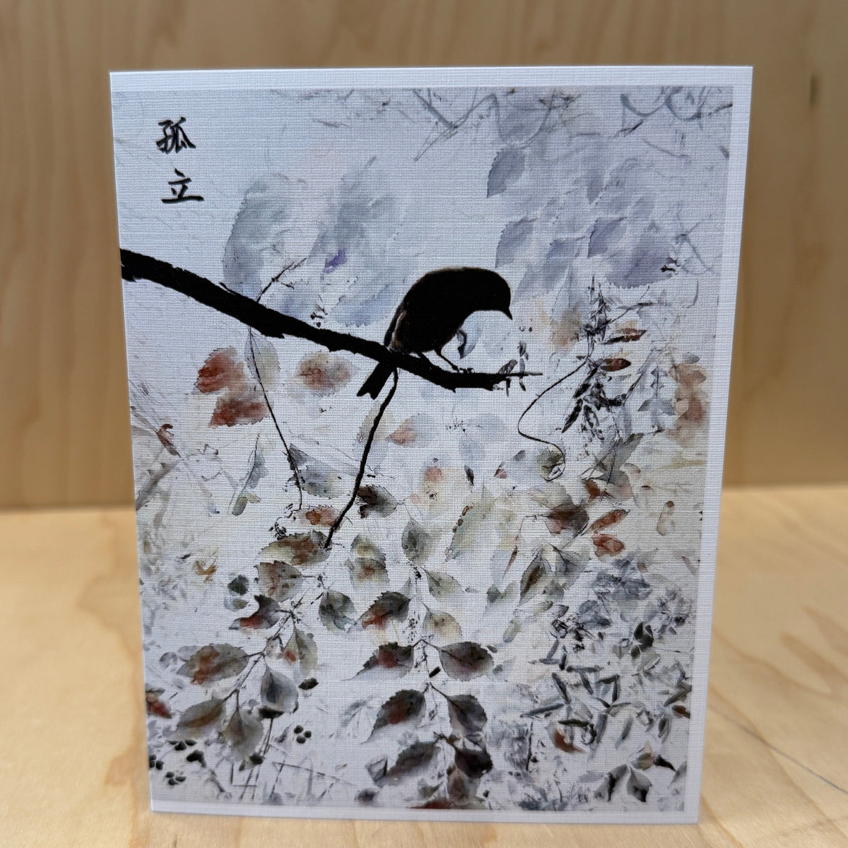 A square art print or card featuring a black bird perched on a branch over a white and gray painted background with subtle brown and red accents, displayed on a light wood surface against a wood-paneled backdrop.