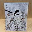 A square art print or card featuring a black bird perched on a branch over a white and gray painted background with subtle brown and red accents, displayed on a light wood surface against a wood-paneled backdrop.